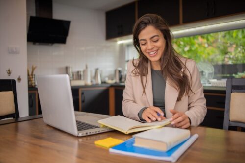 A woman works from home with a notebook and a laptop nearby, a kitchen in the background