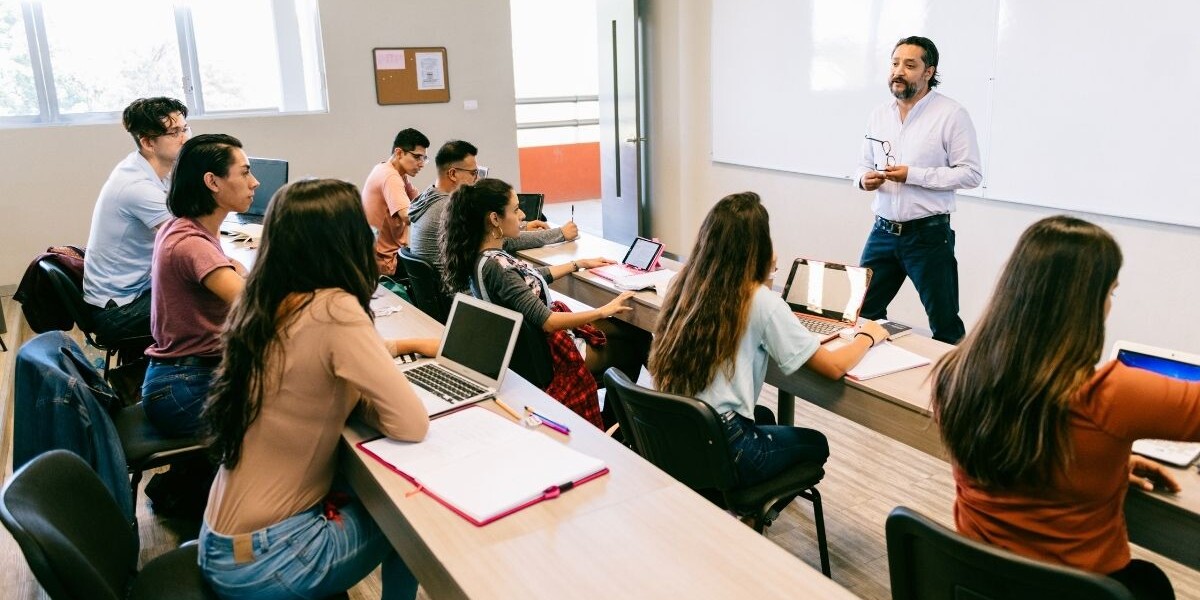 A teacher at the front of a classroom talks to high schools students who are seated